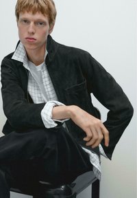 Schwarze Wildlederjacke mit Reißverschluss, getragen über einem weiß-grau karierten Hemd. Das Model hat helles Haar und sitzt entspannt.