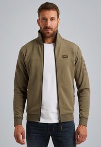 Olijfgroene ritsjack met een gestructureerd patroon, hoge kraag en logo-opdruk, gecombineerd met een wit shirt en donkerblauwe spijkerbroek.