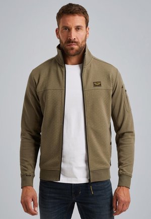 Olijfgroene ritsjack met een gestructureerd patroon, hoge kraag en logo-opdruk, gecombineerd met een wit shirt en donkerblauwe spijkerbroek.