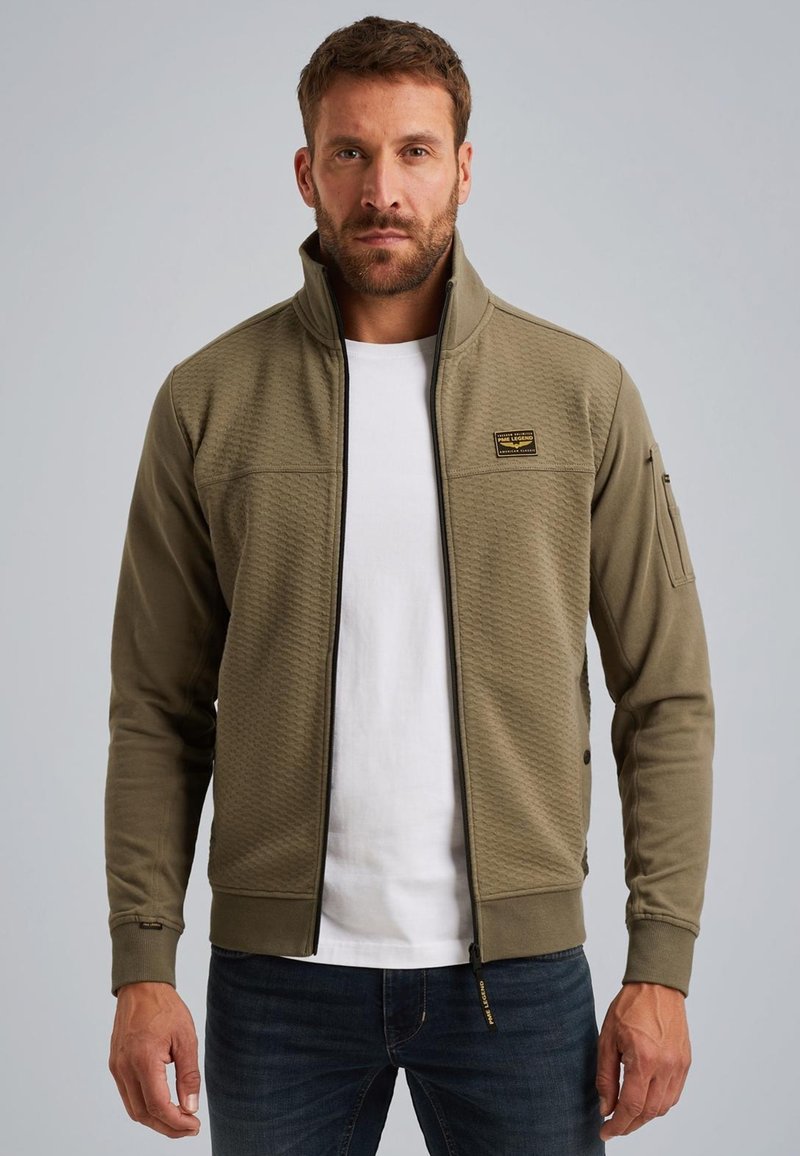 Olijfgroene ritsjack met een gestructureerd patroon, hoge kraag en logo-opdruk, gecombineerd met een wit shirt en donkerblauwe spijkerbroek.