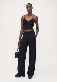 Black cropped knit top with thin straps, paired with high-waisted black wide-leg trousers. Model holds a small black handbag.