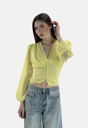 Blusa gialla trasparente con maniche lunghe e voluminose, scollatura a V e dettagli con bottoni sul davanti. Abbinata a jeans azzurri chiari a vita alta.