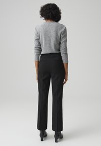 Maglione grigio texture e pantaloni neri su misura. L'outfit è completato da stivali neri con tacco alto. La vista posteriore mostra un design aderente.