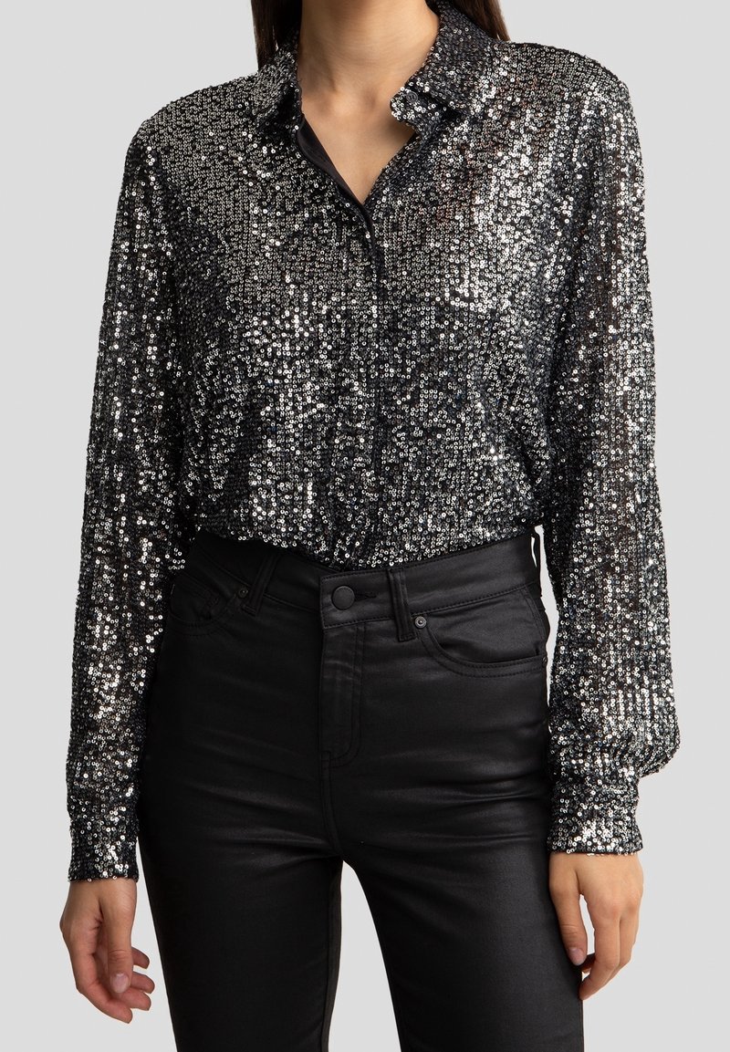 Blouse noire à sequins avec col à boutons. Manches longues et coupe décontractée. Associée à un pantalon noir taille haute.