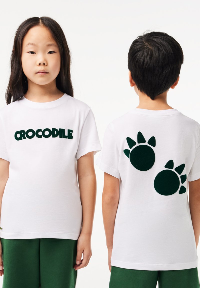 Deux enfants portant des t-shirts blancs et des pantalons verts ; l'avant affiche le texte "CROCODILE", tandis que l'arrière présente deux empreintes de pattes noires.