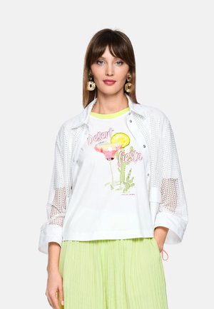 Frau trägt eine weiße perforierte Jacke, ein weißes Grafik-T-Shirt mit der Aufschrift „Desert Fiesta“ mit Cocktail und Kaktus, einen limonengrünen plissierten Rock und goldene Ohrringe.