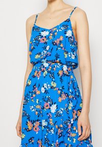 Abito floreale blu con scollo a V, spalline sottili e vita elastica. Il tessuto è leggero con un design fluente.
