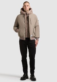 Beige színű puffermellény kapucnival, cipzáros elülső résszel, két oldalsó zsebbel és bordázott mandzsettákkal, egy világosbarna steppelt belső dzseki felett.