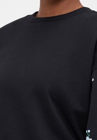 Sweat-shirt noir en coton doux, avec un col rond. Les manches sont ornées d'une broderie florale délicate en bleu clair et blanc.