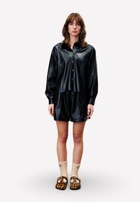 Zwart nepleren overhemd met knopen, gecombineerd met bijpassende korte broek. Model draagt schoenen met luipaardprint en beige gestructureerde sokken.