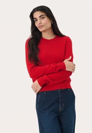 Rode gebreide trui met lange mouwen en een ronde halslijn, gecombineerd met hooggetailleerde blauwe jeans. Zachte textuur met ribbelmanchetten en -zoom.