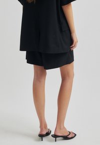 Černý oversized top, odpovídající černé šortky a černé sandály na podpatku. Tkanina vypadá hladce a má uvolněný střih po celém obvodu.