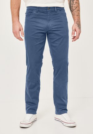 Mann, der blaue gerade geschnittene Hose und weiße Sneakers trägt, steht vor einem einfarbigen hellen Hintergrund.