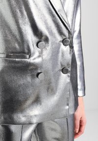 Stříbrný metalický dvouřadý blazer s kulatými knoflíky, hladkou texturou a bočními kapsami; přizpůsobený střih s elegantním designem.