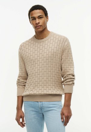 Mand iført beige struktureret striktrøje og lyseblå jeans, stående mod en ensfarvet hvid baggrund med et neutralt udtryk.