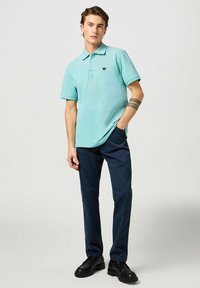 Polo shirt turchese con colletto e maniche corte, abbinato a pantaloni scuri navy e scarpe nere. Presenta un piccolo logo sul petto.
