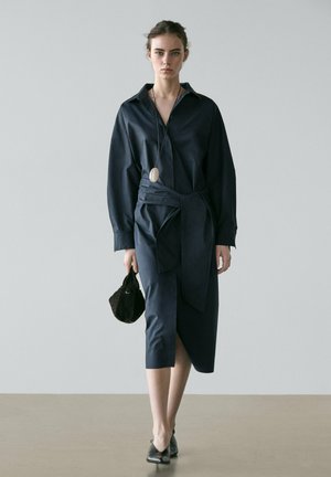 Woman wearing a navy blue belted shirt dress and black shoes, holding a small black handbag, standing against a plain gray background.