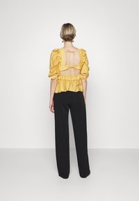 Blusa gialla con volant e maniche a sbuffo, schiena scoperta fissata da lacci, abbinata a pantaloni neri a gamba larga. Texture liscia e colore vivace.