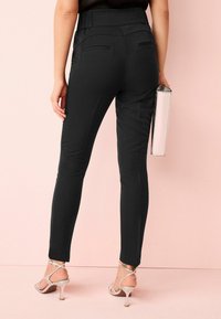 Schwarze Slim-Fit-Hosen mit hoher Taille, mit Gesäßtaschen, kombiniert mit silbernen Riemchen-High Heels und einer metallischen Clutch.