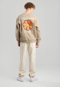 Beige sweatshirt with "CALIFORNIA" text and floral design in orange, paired with light beige pants and white sneakers, shown from the back.