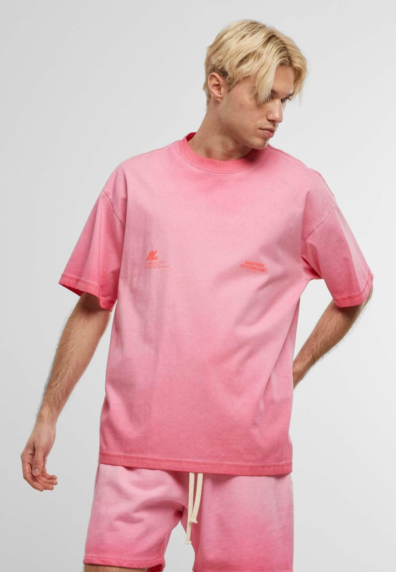 Cămașă oversized de tip t-shirt, de culoare roz cu un gradient, cu mâneci scurte, guler rotund și un mic logo portocalie pe piept.