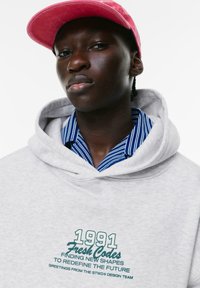 Felpa grigia con tasca a marsupio e testo del logo in verde. Indossata sopra una camicia in cotone a righe blu e bianche. Presenta un cappellino rosa.