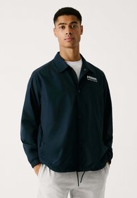 Chaqueta ligera azul marino con cuello, botones a presión, bolsillos laterales y logo de "RBR Red Bull Racing" en el lado izquierdo del pecho. Textura suave.