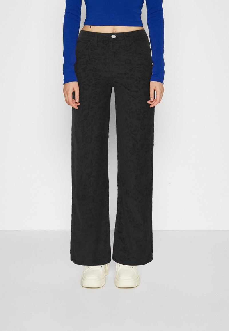 Femme portant un pantalon large en velours côtelé noir, un top bleu à manches longues raccourci et des baskets plateformes blanches, debout sur un fond blanc.