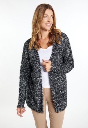 Cardigan fuzzy alb-negru cu un croi deschis, mâneci lungi și o croială lejeră. Purat peste un top alb și pantaloni bej.