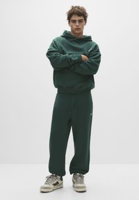 Hanorac verde și pantaloni asortați dintr-un material moale, cu o croială lejeră. Prezintă un cordon la talie și branding subtil pe hanorac.