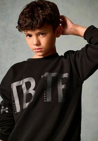 Crni dukserica s hrabrim, odražavajućim printom s natpisima "TB" i "BY TED BAKER" na prednjoj strani. Mekani materijal s ležernim krojem.