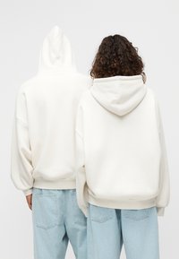 Λευκά φαρδιά φούτερ με καπέλο και χαλαρούς ώμους, με ριμπ μανσέτες και τελείωμα. Φορεμένα με ανοιχτόχρωμα μπλε τζιν, θέα από πίσω.