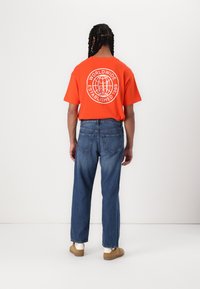 T-shirt orange à manches courtes avec un graphique blanc "WORLDWIDE ESTABLISHED 1969" ; associé à un jean bleu et des chaussures à enfiler marron.