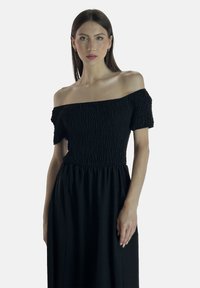 Sort off-shoulder kjole med tekstureret smocket top og flydende skørt, der fremhæver et blødt stof og en afslappet silhuet.