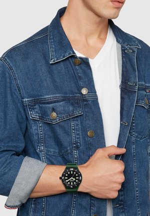 Man wearing a blue denim jacket over a white V-neck shirt, showing a green and black analog wristwatch on his left wrist.
