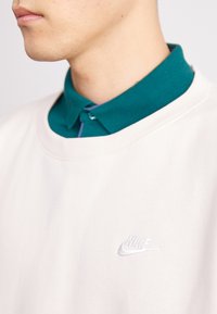Sānu profils cilvēkam, kurš valkā tirkīza krāsas krekls ar apkakli zem baltas Nike treniņjakas, uz kura krūšu daļas ir neliels izšūts logs.