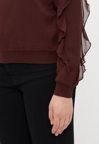 Top bordeaux com detalhes em tecido transparente e franzido nas mangas. O corpo é sólido com punhos canelados. Usado com jeans pretos.