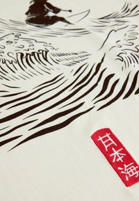 Bavlnená tkanina s čierno-bielym vlnovým vzorom s surferom, doplnená červenou vyšitou visačkou s japonskými znakmi.