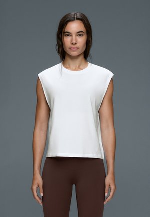 Femme aux longs cheveux foncés portant un haut blanc sans manches et des leggings marron, debout devant un fond gris uni.