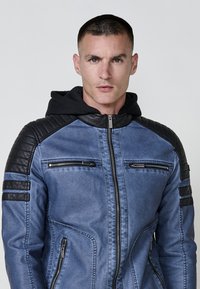 Giovane uomo con capelli corti, che indossa una giacca blu e nera con zip, spalle trapuntate e cappuccio, in piedi davanti a uno sfondo chiaro e uniforme.