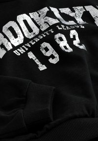 Sweatshirt noir avec impression texturée blanche affichant "BROOKLYN", "UNIVERSITY" et "1982". Comprend des poignets côtelés et une texture de tissu douce.