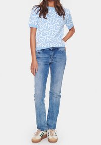 Žena v světle modrém vzorovaném krátkém svetr, středně opraných rovně střižených džínách a bílých teniskách se zelenými pruhy, stojí se rukou v kapse.