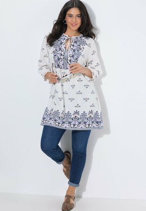 Kvinde iført en hvid tunika med blå blomsterprint, der holder briller, kombineret med opkørte blå jeans og brune sandaler.