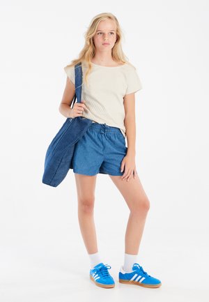 Ležérní outfit složený z krémové strukturované košile, modrých džínových šortek a modrých tenisek s bílými pruhy; džínová crossbody taška.