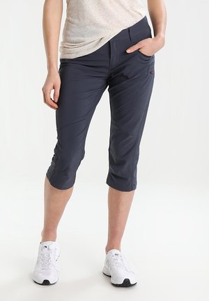 Femme portant un pantalon court gris foncé et des baskets blanches, debout avec une main dans la poche sur un fond blanc uni.