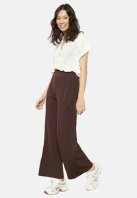 Brown wide-leg pants with a high waistband, paired with a short-sleeved white blouse featuring embroidered details, worn with white sneakers.