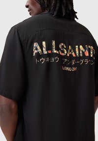 Czarna koszulka z krótkim rękawem z wielokolorowym logo "ALLSAINTS" na plecach z fakturowanym wzorem węża, tworząca odważny design.