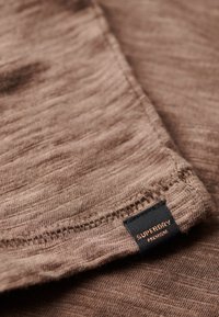 Bruine, gestructureerde stof met een zachte afwerking, met een zwart label met de tekst "SUPERDRY PREMIUM" aan de rand gestikt.