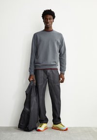 Sweatshirt gris à col rond, pantalon gris foncé et baskets jaunes avec accents rouges et bleus. Modèle tenant une veste assortie.