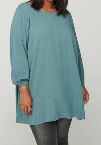 Haut ample couleur sarcelle avec un col rond et des manches trois-quarts. Le tissu présente un motif texturé subtil. L'ourlet est incurvé.